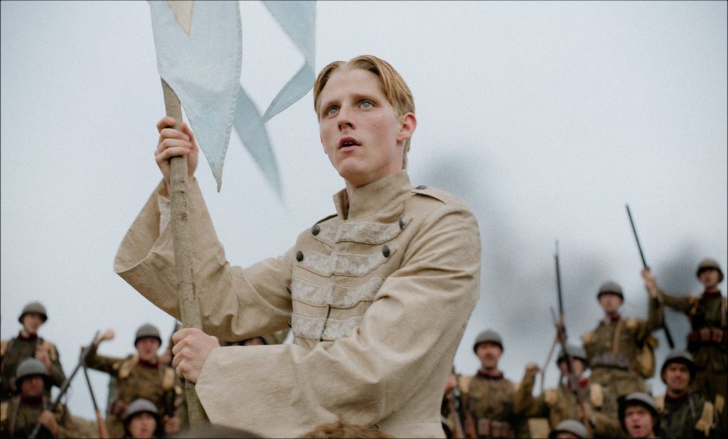 Un jeune homme en costume de scène brandit un drapeau devant des soldats dans Coward