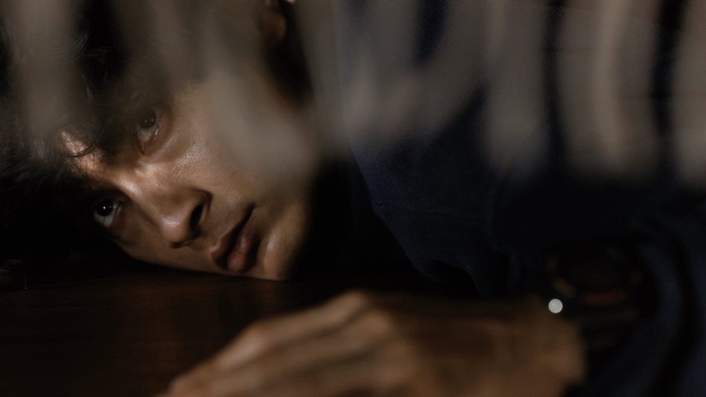 Un homme, la tête posée sur une table, regarde fixement dans la pénombre, dans Under Your Bed de Mari Asato